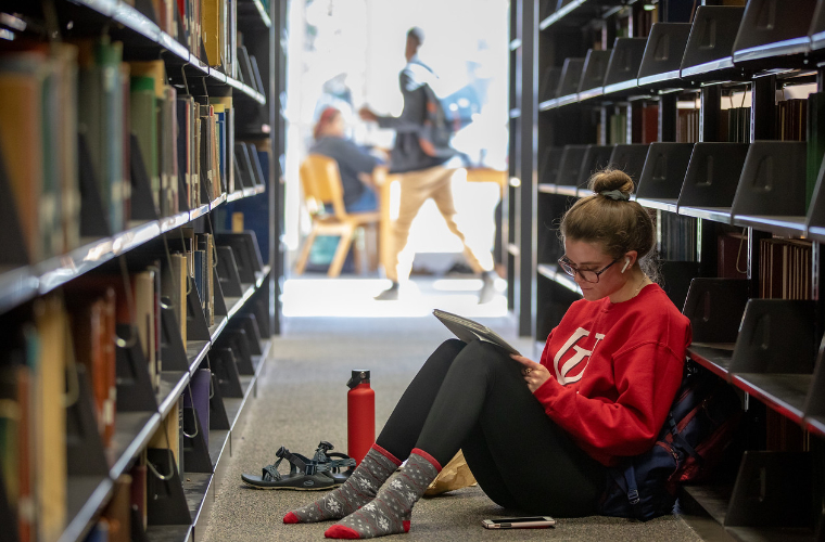 UL Lafayette student sits shoeless on library floor between shelves writing