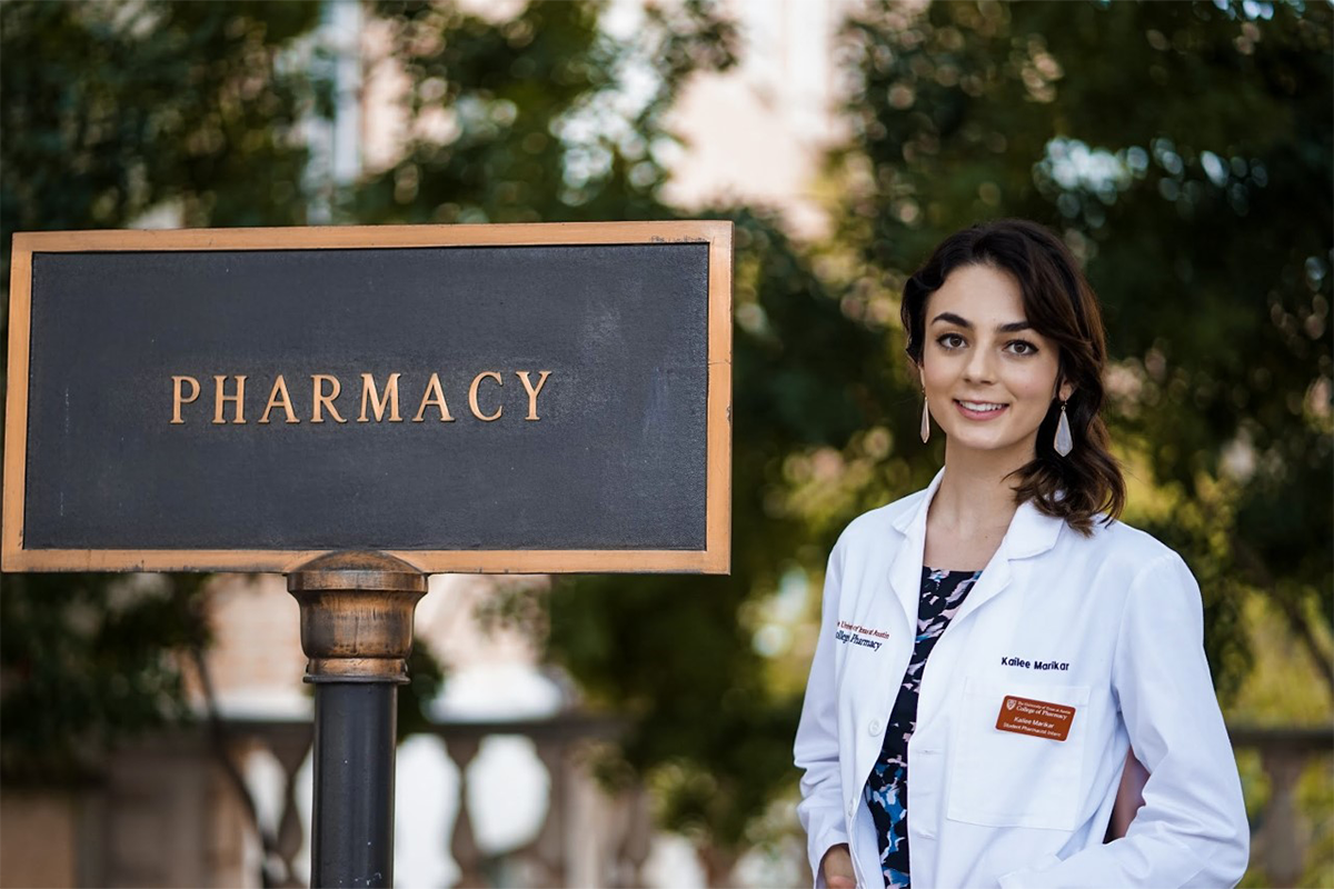 Bachelor of General Studies alumni Kailee Marikar stands in a white lab coat near a "pharmacy" sign shortly before earning her doctor of pharmacy degree.