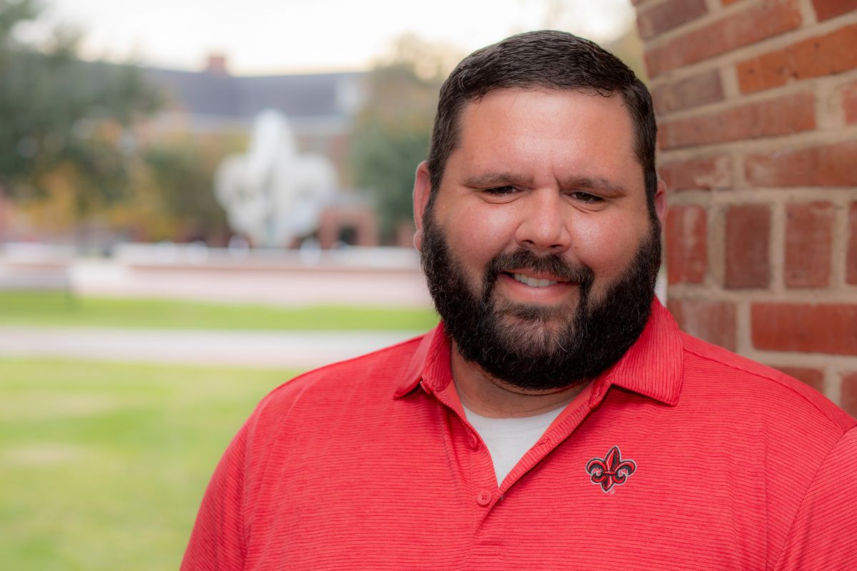 Zach Badon, wearing a red UL Lafayette polo, poses for a photo in the University's quad. He earned his BSBA in Management online in 2021 and his MBA online in 2023.