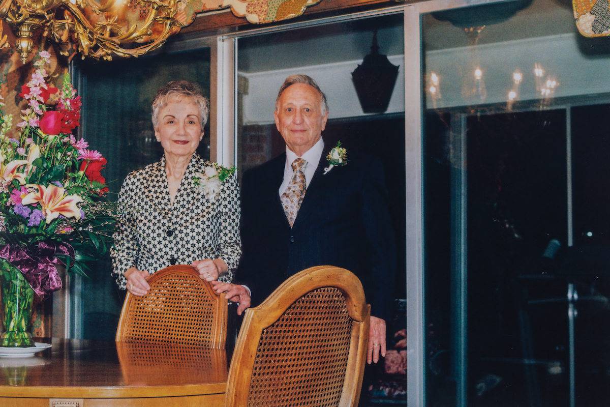 A couple stands next to a bouquet of flowers sitting on a wooden table.