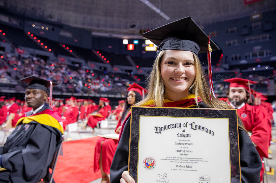 MS in Informatics graduate wearing a black cap and gown smiles with diploma at UL Lafayette commencement ceremony