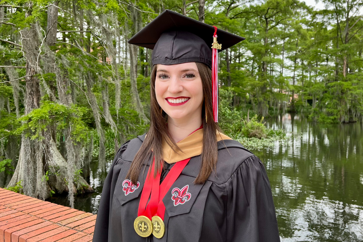 Kaley Carroll, wearing her graduation cap and gown, smiles for a photo in front of Cypress Lake on UL Lafayette's campus.