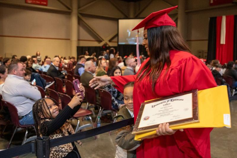 Halle Castille, holding her diploma after walking off the graduation stage, hugs her son, age 7, while her daughter, age 4, takes a photo.