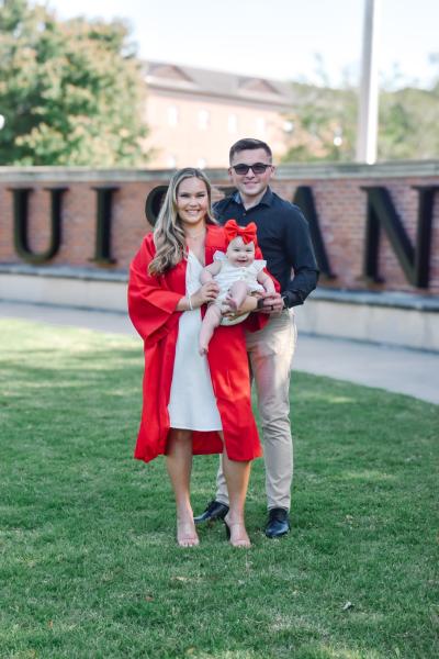 Online general studies grad, in her UL Lafayette graduation gown, poses for photo with husband and their baby