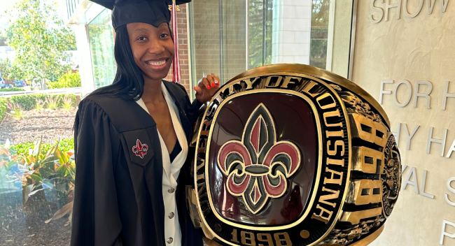 Wearing her cap and gown, April Piper stands next to a large replica of UL Lafayette's class ring.