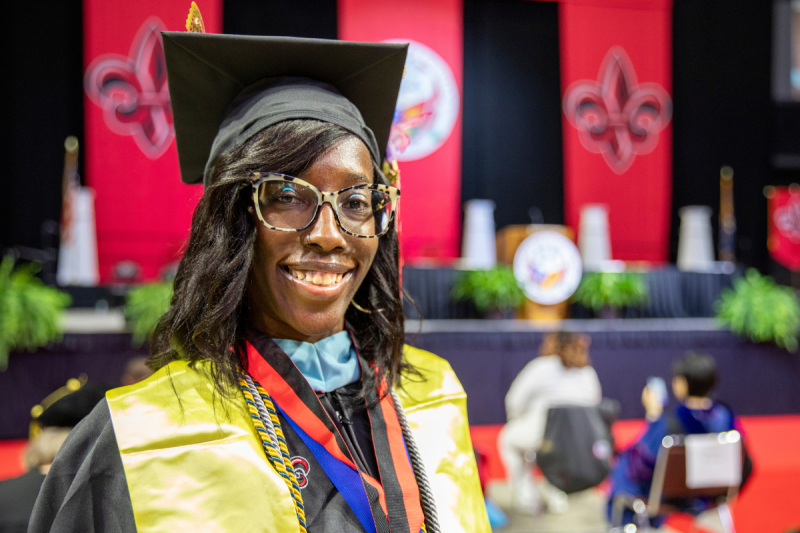 Denise Mallard, wearing her graduation cap and gown, smiles for a photo in front of the stage at Commencement.